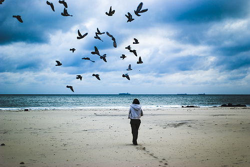 A woman on a beach with footbprints behind her and a flock of birds in the sky
